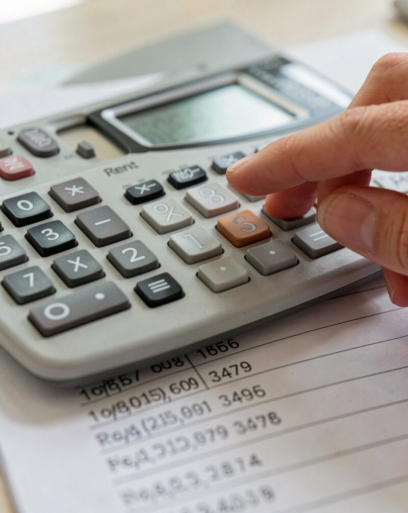 A household budget worksheet on a kitchen table with a small calculator, rent receipts and utility bills in two piles, a mug of tea